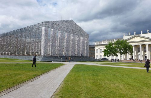 Die Installation der argentinischen Künstlerin Marta Minujin „The Parthenon of Books“ zählt ohne Frage zu den sichtbarsten Ausstellungsstücken der diesjährigen documenta – und gilt als Highlight der Kasseler Ausstellung. 50.000 Bücher winden sich in bis zu 14 Meter Höhe um ein Metallgerüst, das einen maßstabsgetreuen Nachbau des griechischen Vorbilds in Athen darstellt. Zweifelsfrei ist die Installation damit einer der aufwendigsten Werke der Kunstschau. Bereits im Oktober 2016 startete das documenta-Team einen Spendenaufruf auf der Frankfurter Buchmesse: Privatleute und Verlage wurden aufgefordert, Bücher mit einer besonderen Geschichte zu spenden, nämlich solche, die ehemals verboten waren oder es noch immer sind. Damit ist das Kunstwerk eine Bild gewordene Bibliothek verbotener Bücher: „Marta Minujins Projekt sprengt allerdings die Dimensionen dieser Bibliotheken: Denn die Installation steht pars pro toto für alle jemals verbotenen Bücher weltweit und wird damit zu einer Art Mahnmal, einem temporären Memorial für die Freiheit der Gedanken und Weltsichten. Das ,Parthenon of Books‘ beweist vor allem, dass das kollektive Gedächtnis der Menschheit stärker ist als alle Verbote und Zensurmaßnahmen“, lobt so etwa Deutschlandfunk Kultur. 