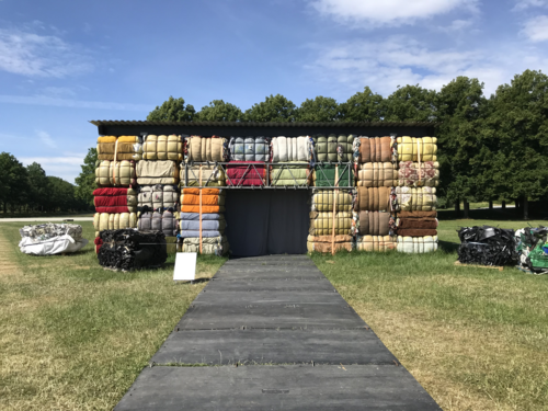 Das interdisziplinäre Künstler:innenkollektiv The Nest Collective aus Nairobi hat für Kassel die Installation „Return to Sender – Delivery Details“ entwickelt, in der Experten über die fatalen Folgen von Secondhand-Kleiderspenden in afrikanische Länder berichten. Die Videoinstallation ist in einer Behausung aus Kleiderballen zu sehen, wie sie in tausenden Tonnen nach Afrika verschifft werden. 40 Prozent dieser Ware ist unbrauchbar und landet dort direkt auf dem Müll.